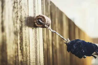A gloved hand holding a paint roller staining a fence.