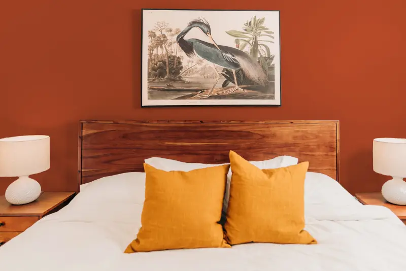 Close-up of bed with wood headboard, white sheets, and a bright rusty orange painted wall behind it.