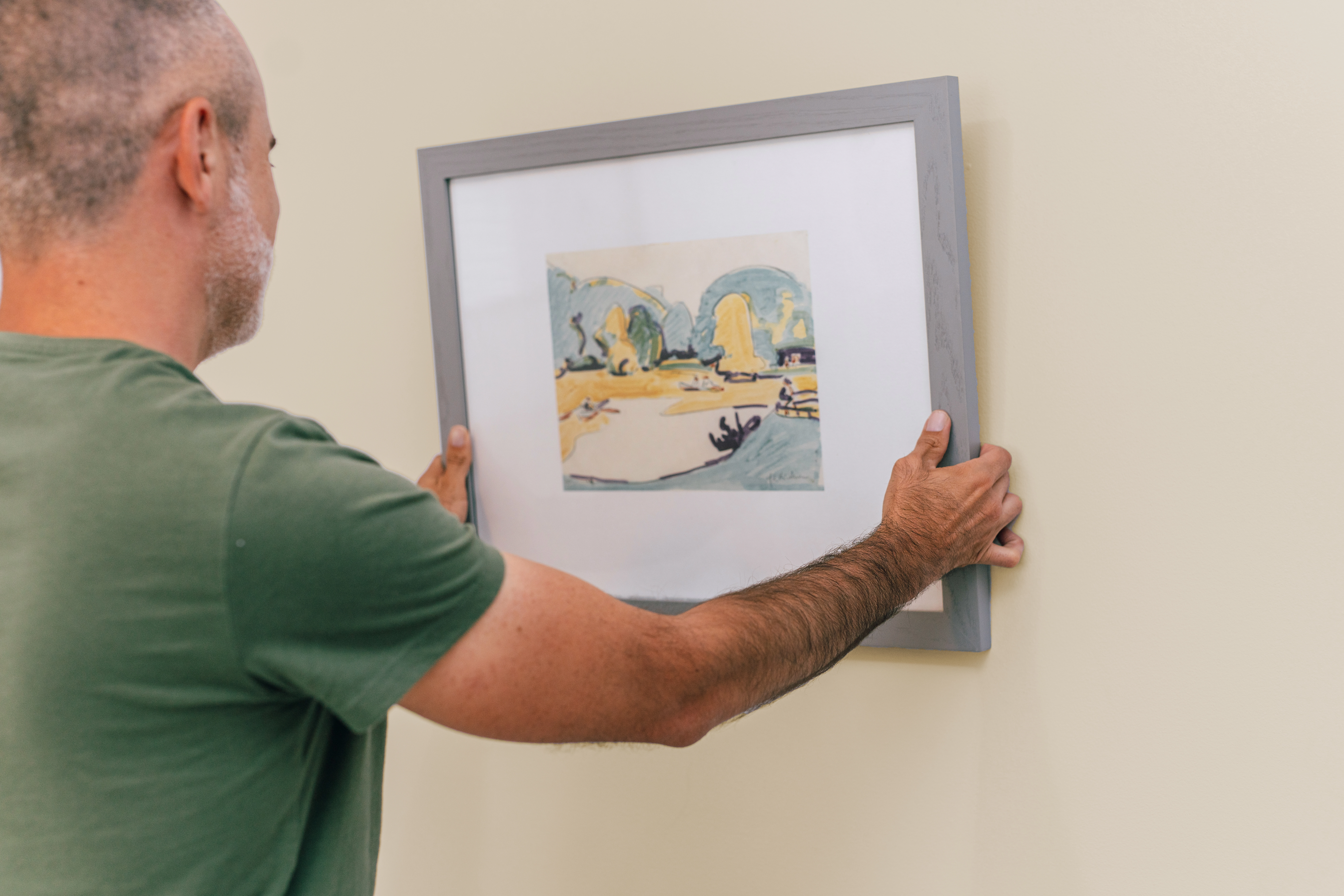 Behind shot of man holding up a gray framed painting and positioning it to hang on a wall.