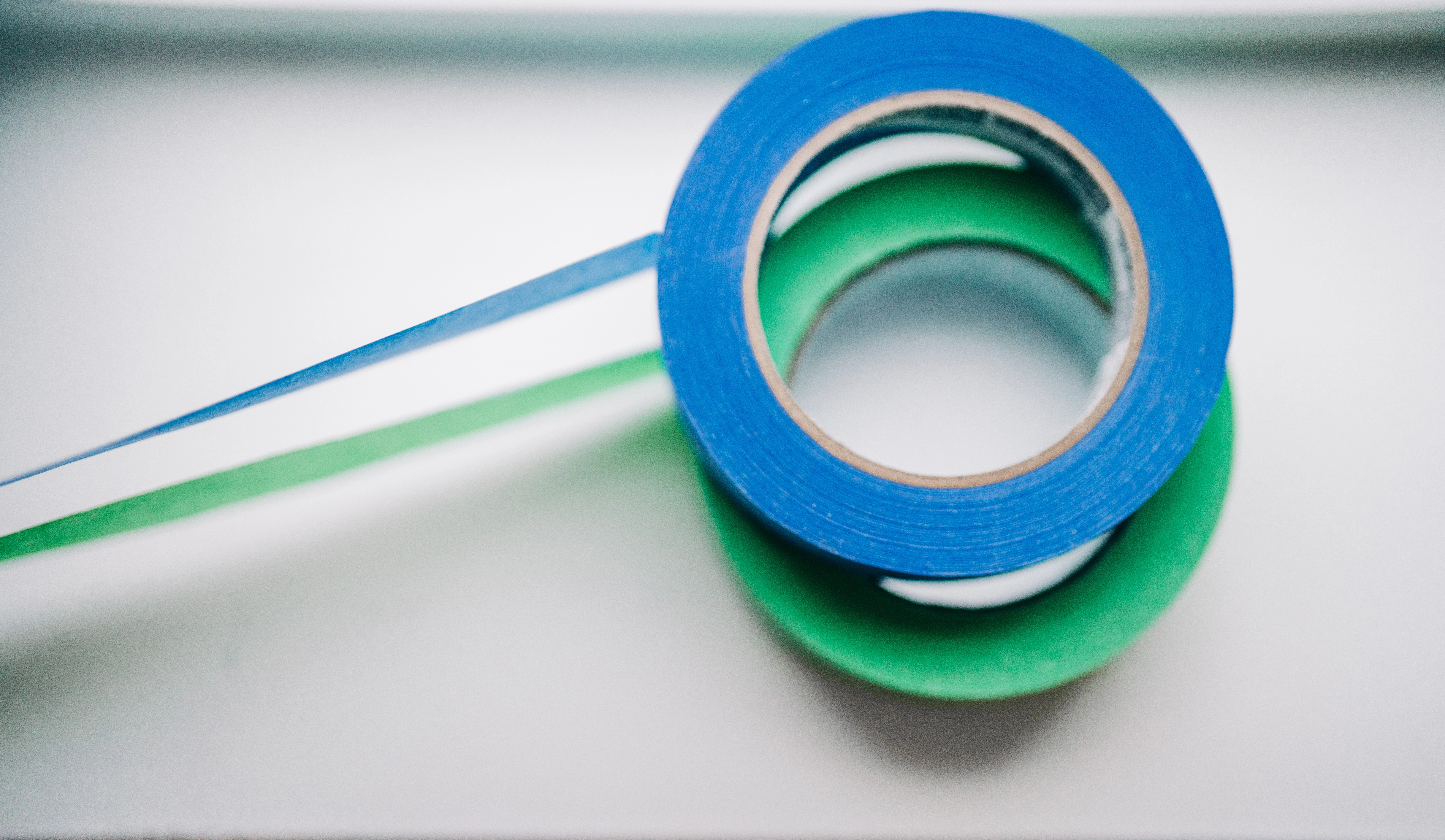 A roll of blue painter's tape stacked on green painter's tape. Both have pieces of tape unrolled.