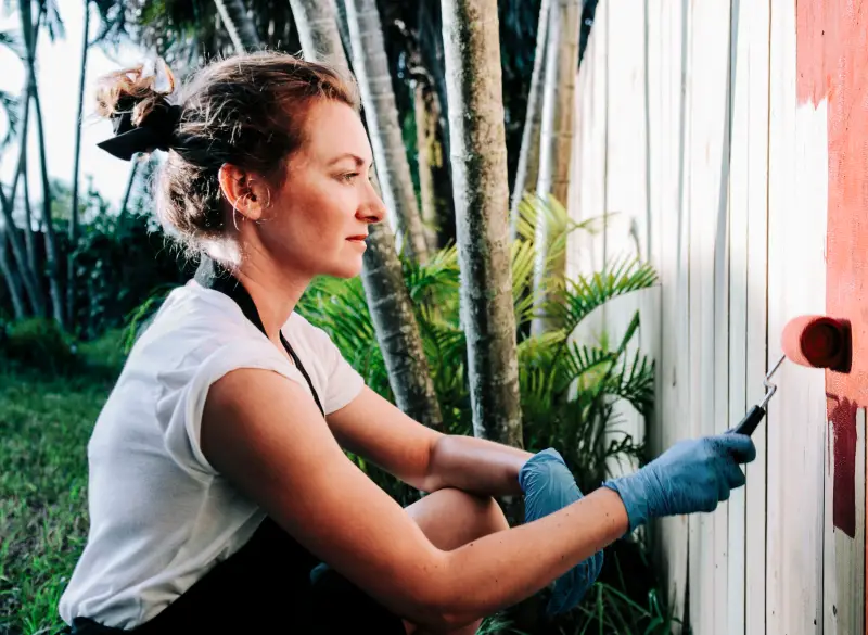 Close-up of woman crouched in yard, using paint roller to paint a white wood fence a peachy red.