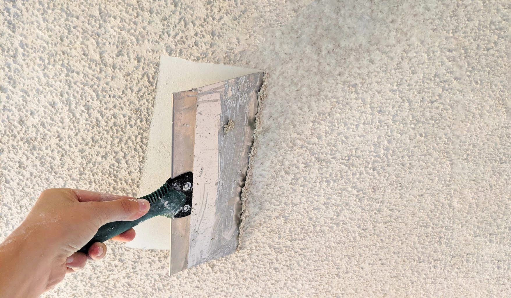 Close-up of someone using a paint scraper tool to remove white painted popcorn ceiling.