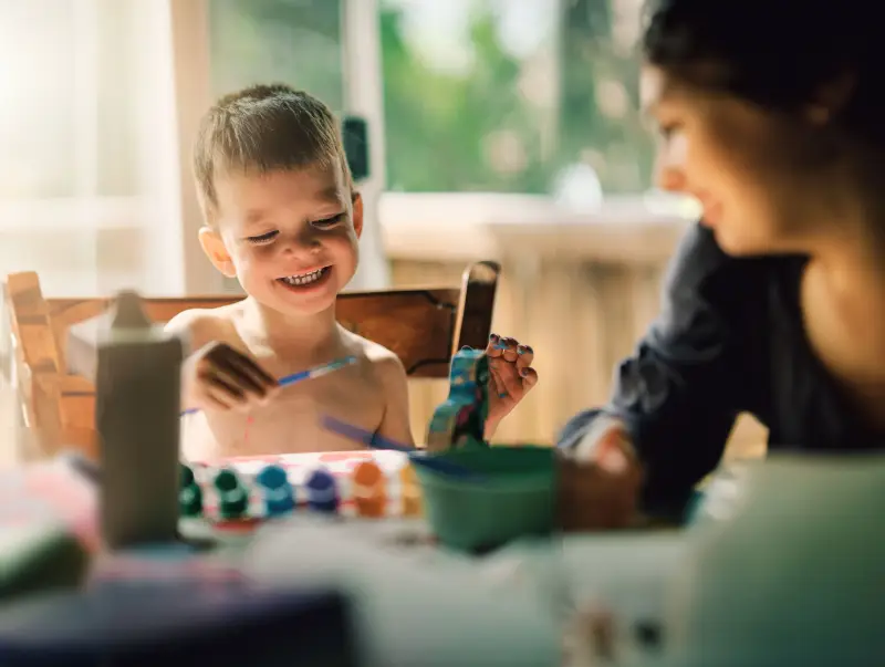 Focused shot of child laughing and holding a paint brush above painting. His mother sits beside him.