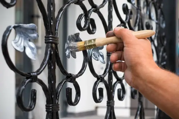 Hand holding a paintbrush and painting an ornate metal gate with silver paint.