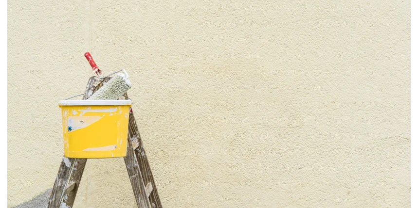 A paint roller sits in a bucket on top of a ladder in front on a painted exterior wall