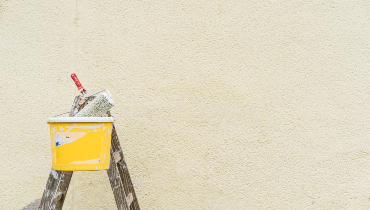 A paint roller sits in a bucket on top of a ladder in front on a painted exterior wall