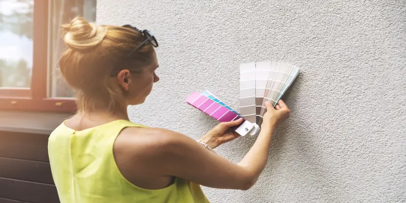 Female holding paint samples up to the side of a house exterior made of stucco to select exterior paint colors.