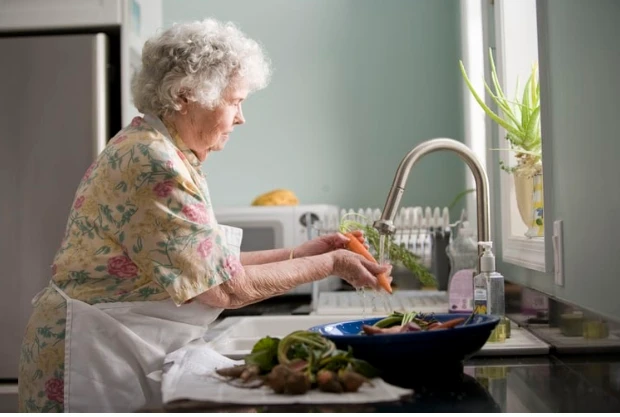 An older women wearing a floral dress and an apron stands at a kitchen sink and washes a carrot