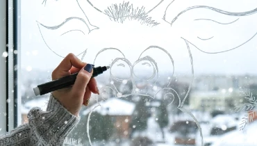 Woman using white paint marker to paint holiday deer on window with snow falling outside.