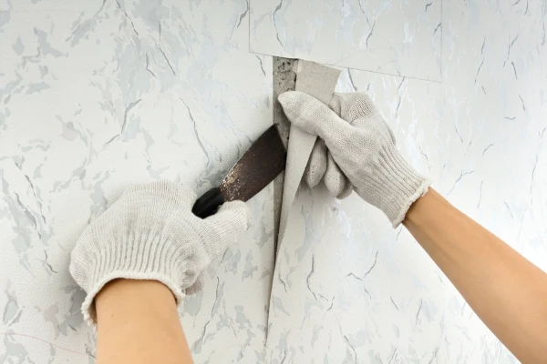A worker wearing grey gloves and holding a putty tool removes old wallpaper from the wall in preparation for painting.