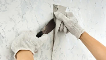 A worker wearing grey gloves and holding a putty tool removes old wallpaper from the wall in preparation for painting.