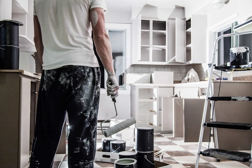 Photo of a paint-covered man holding a used roller brush looking at a kitchen being painted white