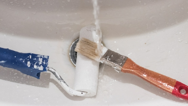 Paint roller and brush being cleaned in a sink under running water.