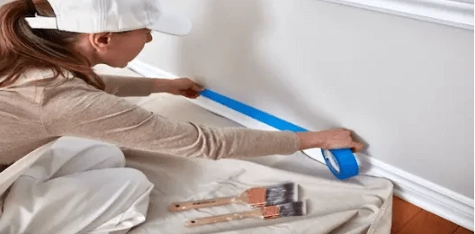 Woman in hat and overalls using painters’ tape to prep wall for painting.