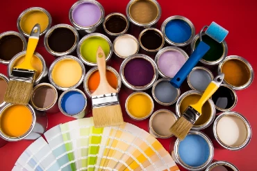 Paint brushes and a small roller sitting on top of open container of different color paint, on a red background.