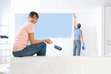 Smiling woman holding a paint-roller in her hand while her husband is applying blue paint to the living room wall using a paint brush.