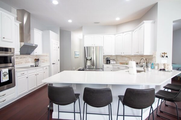 Modern kitchen with white cabinets and large white countertop with black chairs.