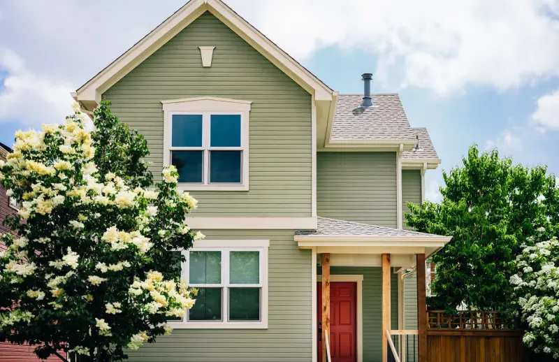 Home with light green wood siding, white trim, dark red front door, stained wood connected fence.