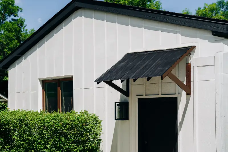 White painted wood garage with stained wood trim, black metal roofing and door, and shrub near door.