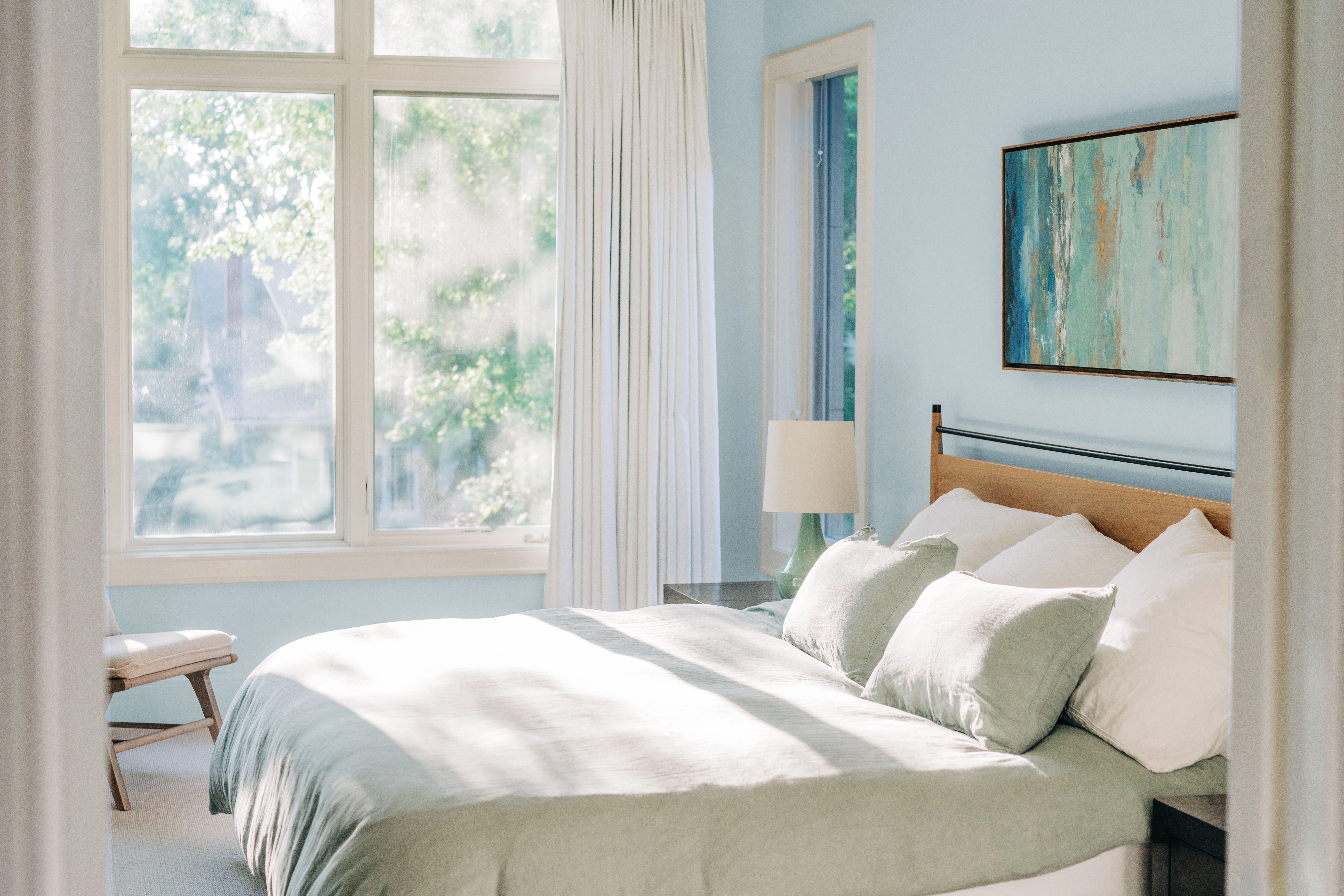 Sunny, light blue bedroom and soft white trim. There's a large window, bed, side tables, and chair.