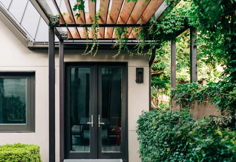 Wood slotted back patio cover with privacy wall and overgrown plants. Black painted door and trim.