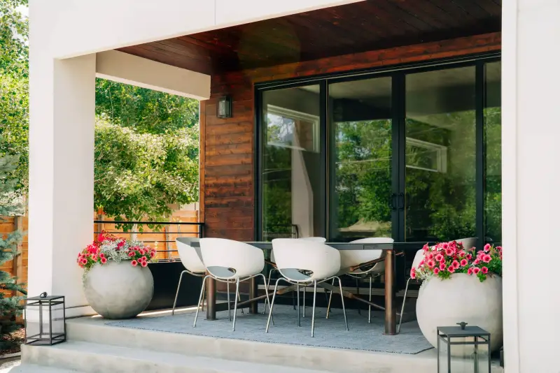 Cement patio and stained wood siding with black trim windows. There's a table, chairs, and flowers.