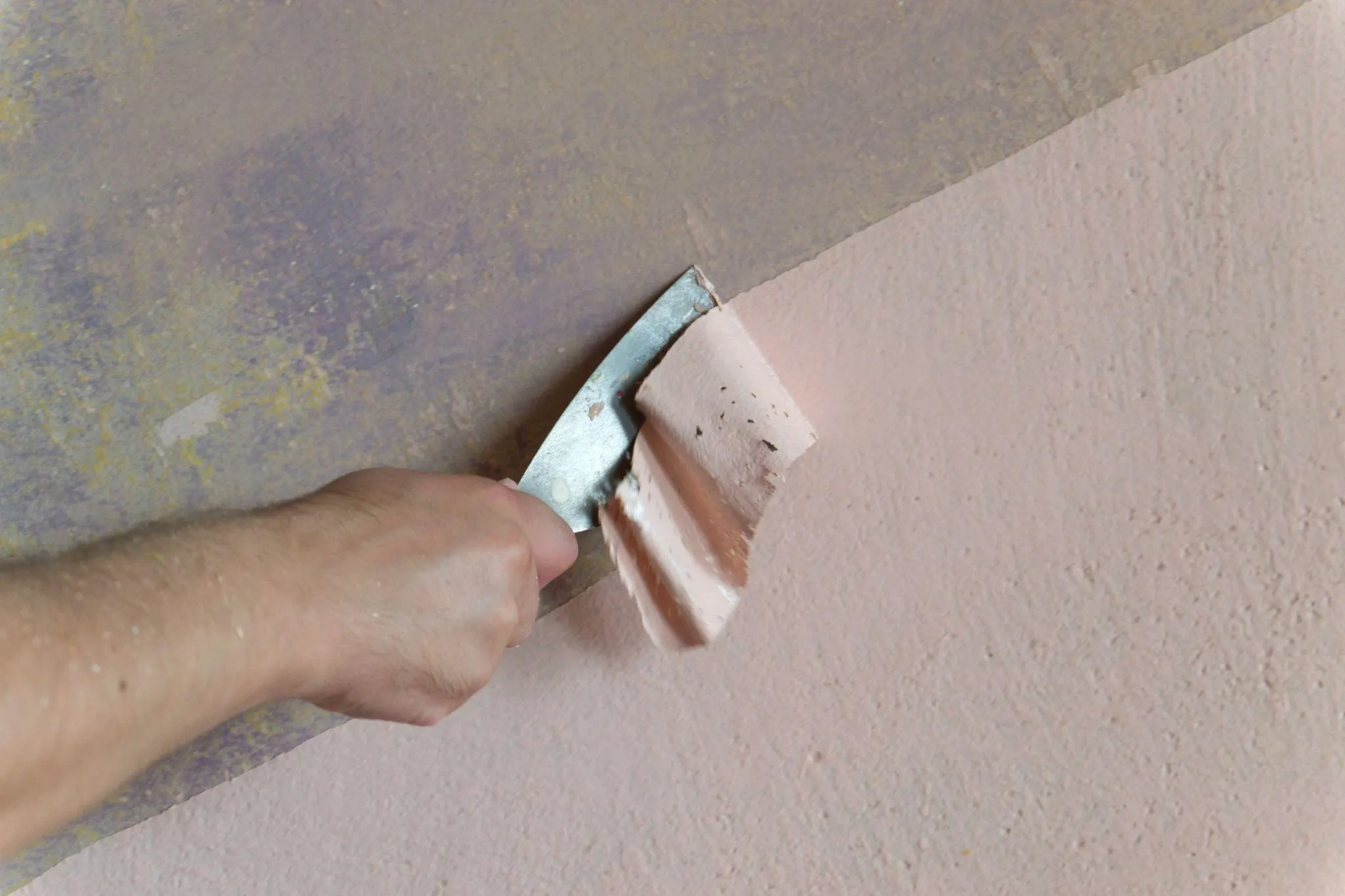 Someone’s hand using a scraper to remove paint from a concrete wall.