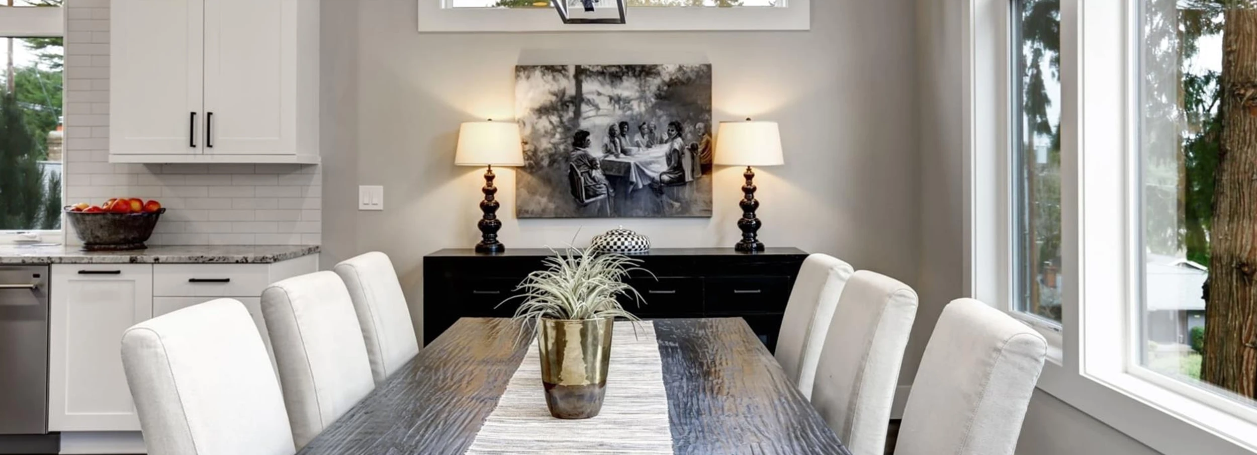 Dining table decorated with woven table runner and potted plant beside white chairs in home dining room with gray walls.