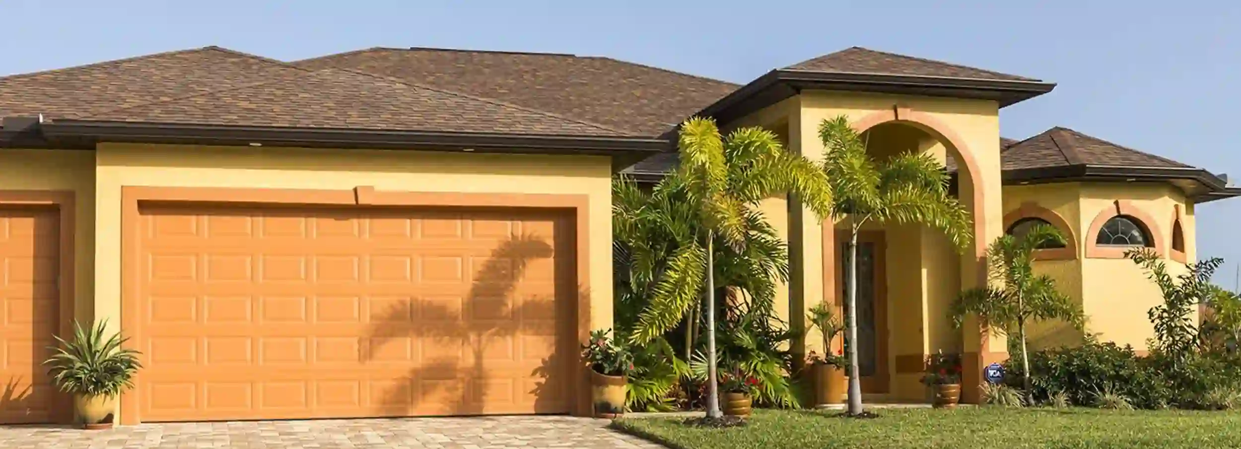 Sunny Florida home with two car garage and entrance surrounded by small palm trees.