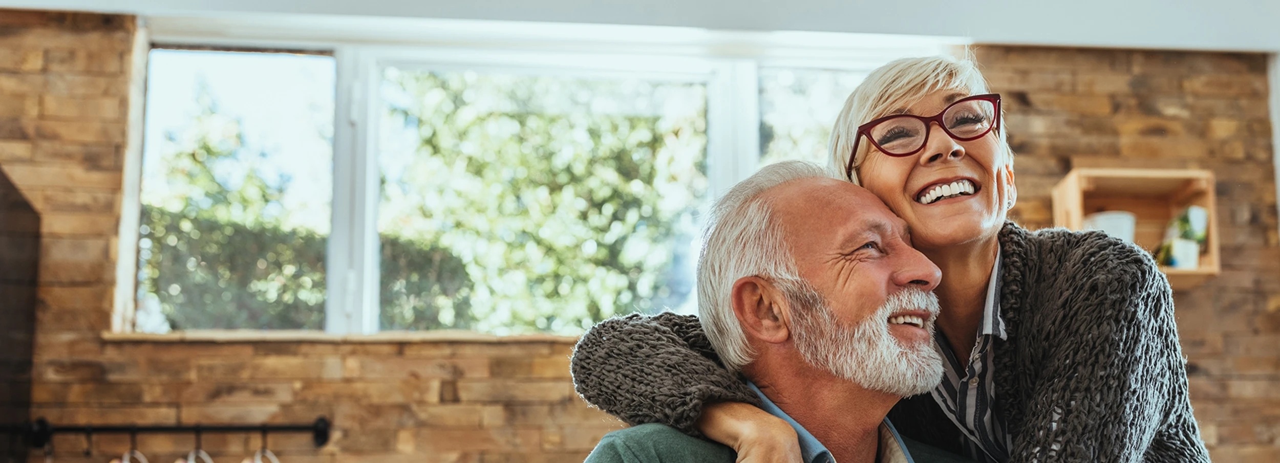 Senior couple smiling and embracing in front of large window set in wood panel wall inside cozy home.