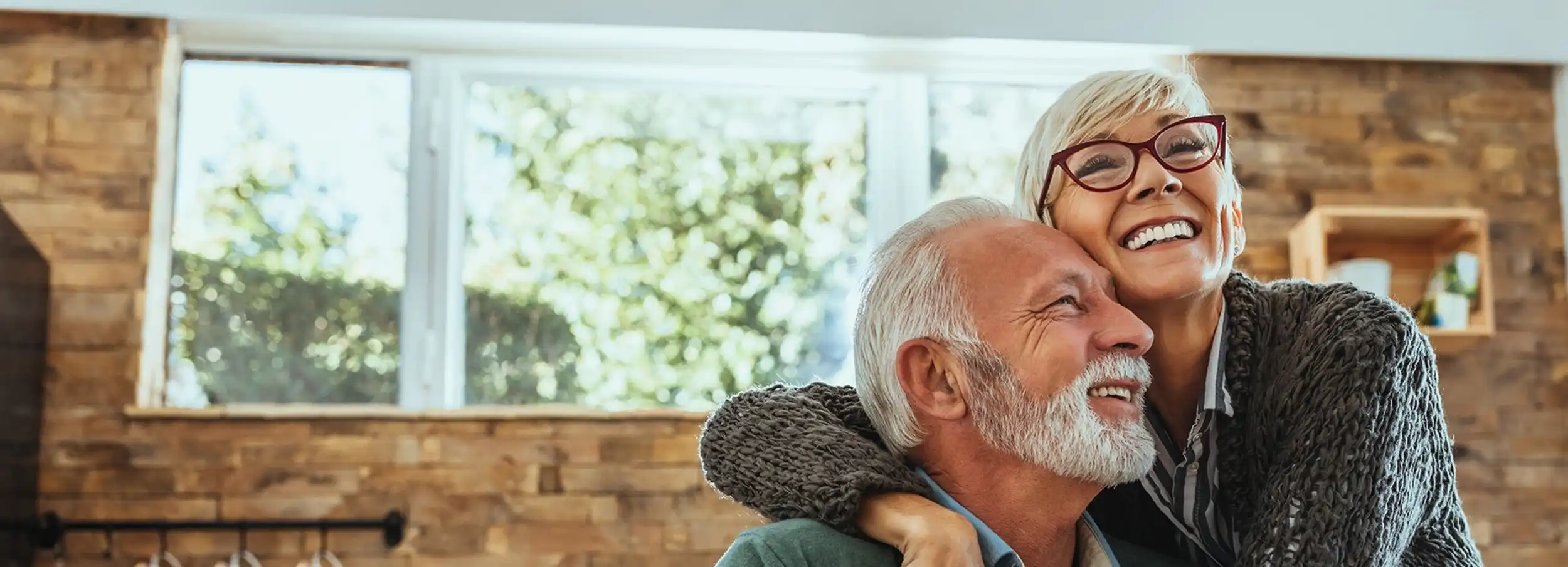 An older man and woman hugging with a brick fireplace in the background.