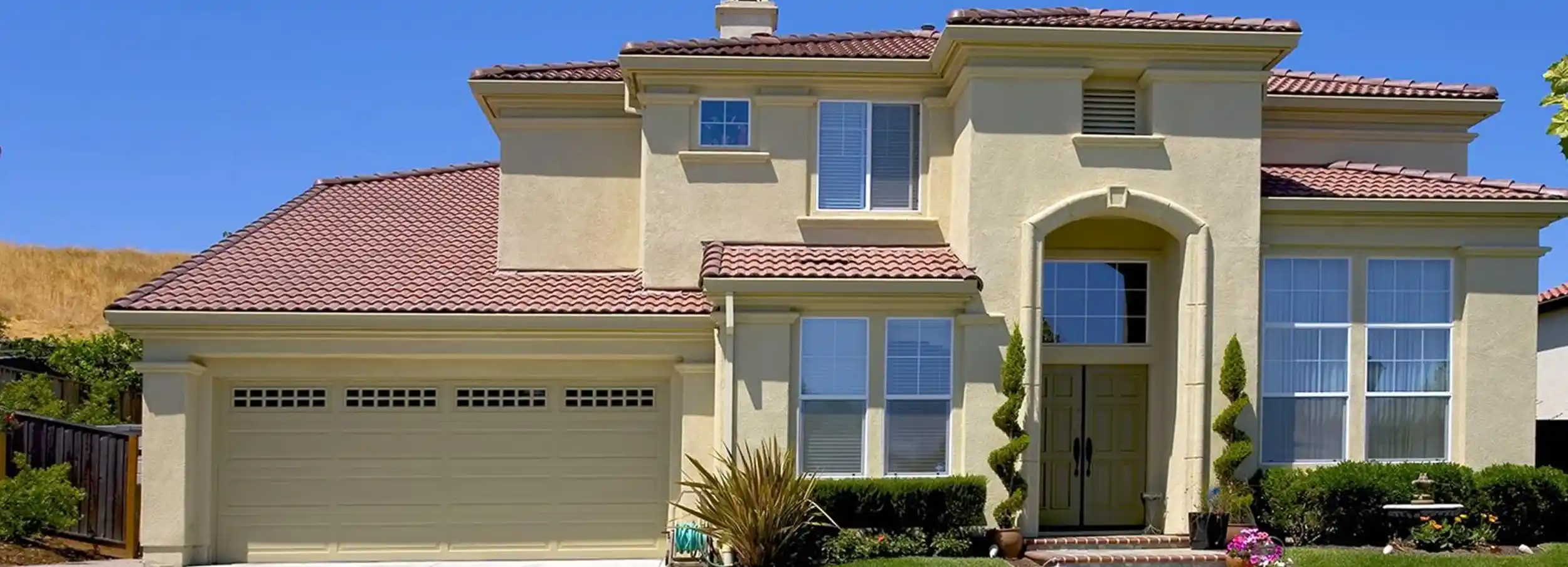 Two story southwestern home with tiled roof and garage and pair of topiary bushes beside entrance.