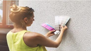 Female holding paint samples up to the side of a house exterior made of stucco to select exterior paint colors.