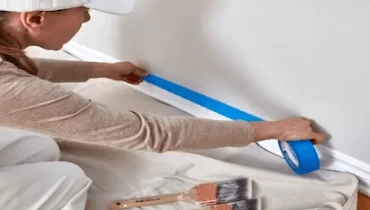 Woman in hat and overalls using painters’ tape to prep wall for painting.