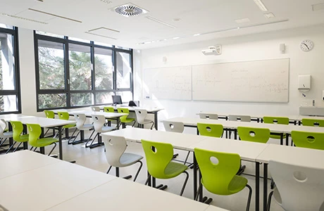 The interior of an empty classroom with white walls, long white tables and white and lime green chairs. 