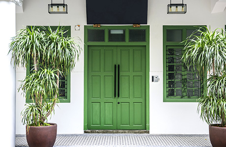 The pristine front entrance of a white and green home with green doors and two plants in front.